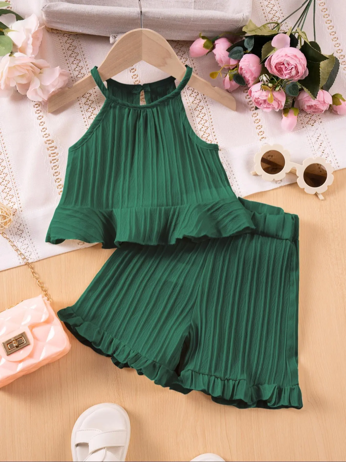 A two-piece children's outfit set in forest green, featuring a sleeveless halter-style top and matching shorts with pleated detailing and ruffle trim, styled on a wooden hanger over a white lace cloth with pink flowers, sunglasses, a small pink purse, and white shoes.
