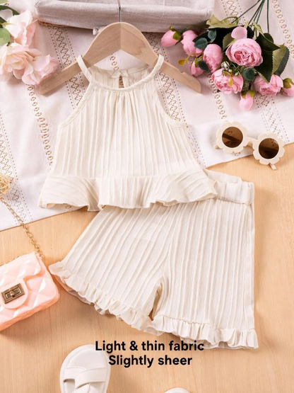 A two-piece baby girl's outfit set featuring a cream-colored pleated halter top and matching ruffled shorts, displayed on a wooden hanger with decorative props including pink flowers, sunglasses, and a small purse.