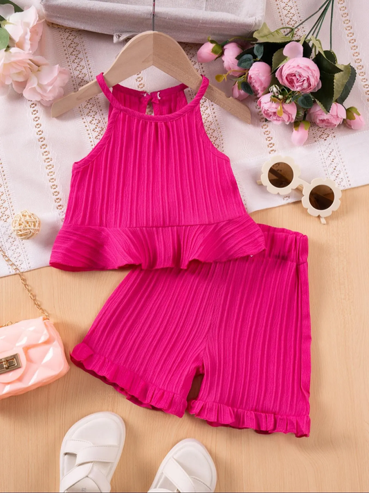 A bright pink two-piece children's outfit set with a sleeveless ruffled top and matching ruffled shorts, styled in a flat lay with flowers, sunglasses, a small bag, and white sandals on a light wood surface.